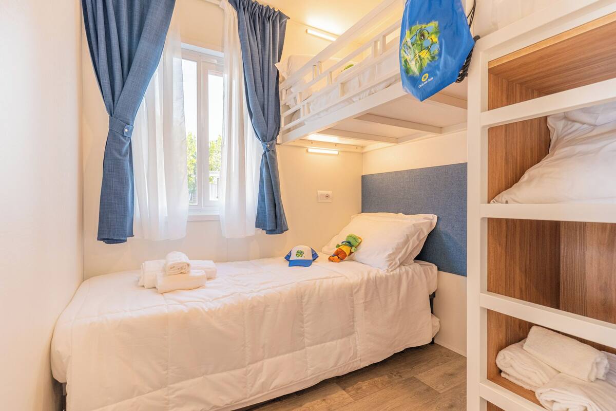 Une chambre avec un lit superposé, une fenêtre, un lit avec un oreiller et un jouet, et une serviette sur le sol.