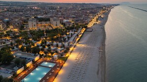 Un paisaje urbano costero con un hotel frente a la playa que cuenta con piscina, tumbonas y un paseo marítimo.