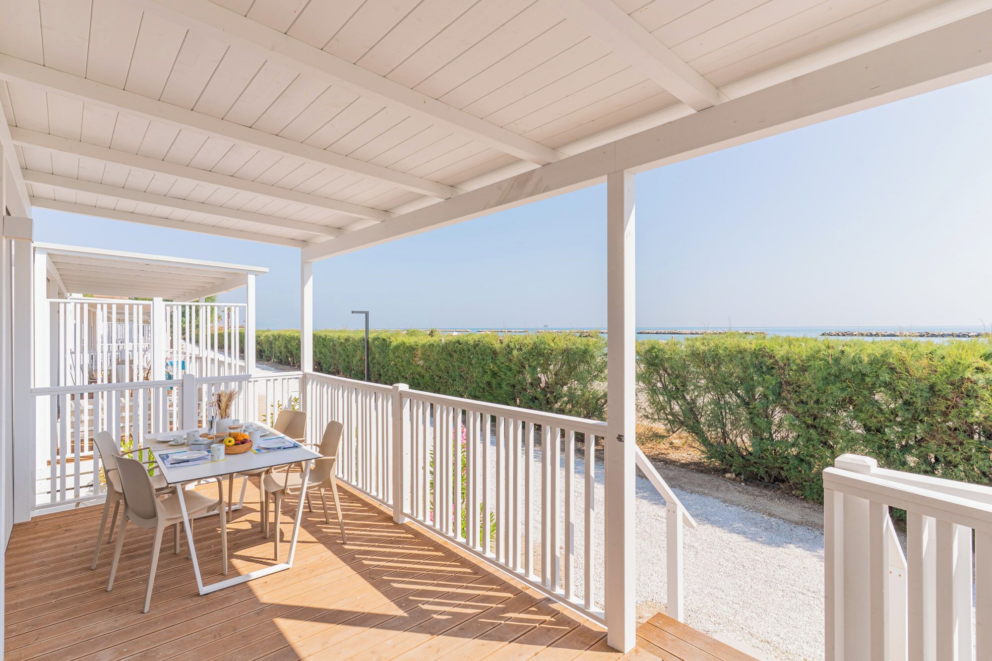 A covered outdoor area with a table and chairs, a white railing, and a view of the sea and greenery.
