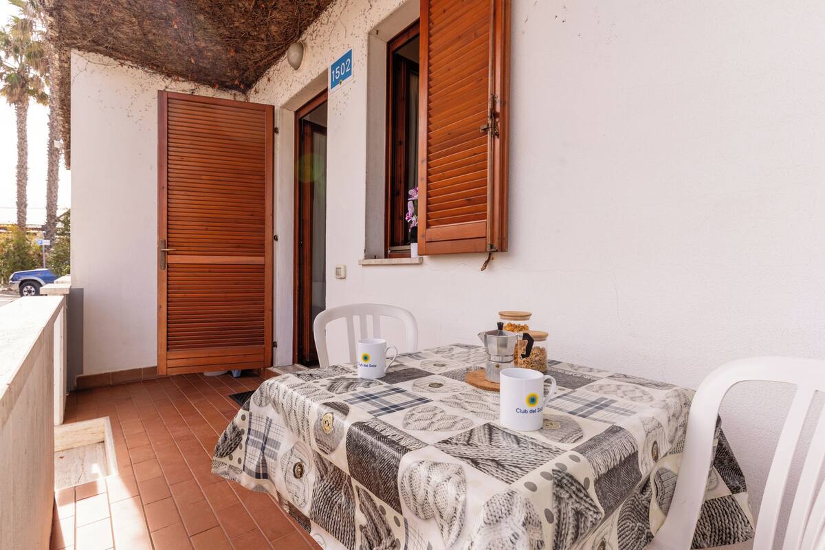 Un balcon avec une table recouverte d’une nappe à motifs, deux chaises blanches et une porte en bois sur laquelle est accrochée une enseigne.