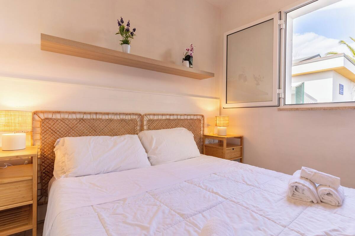 Une chambre avec un grand lit, deux tables de chevet, une commode, une fenêtre et une étagère murale décorée de fleurs.