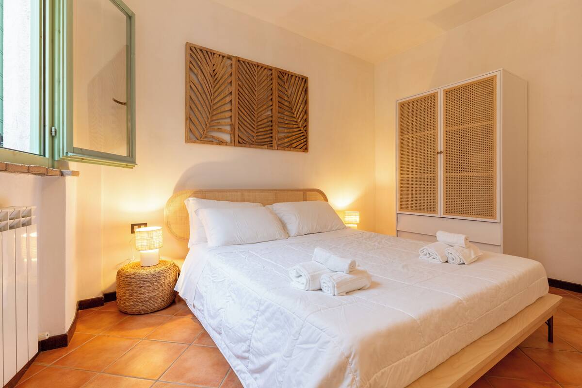 Un lit bien fait, avec du linge de lit blanc et des serviettes pliées, une lampe de chevet, une petite table d’appoint en osier et une grande armoire à portes à persiennes.