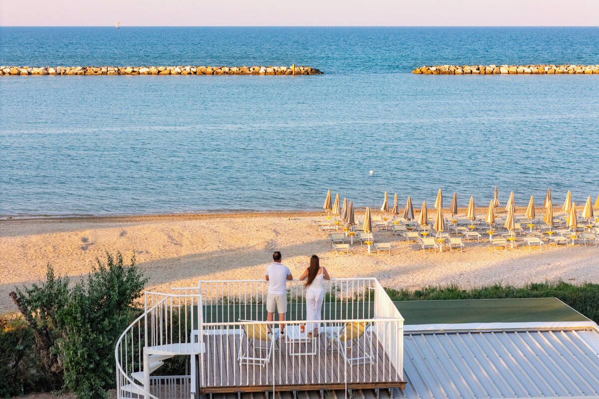 Une vue sur la plage avec une piscine, un couple debout sur une terrasse et une rangée de chaises de plage.