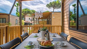 Una terraza de madera con una mesa dispuesta para el desayuno, incluyendo croissants y café, con vistas a una zona residencial con casas y árboles.
