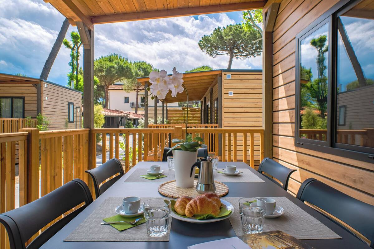 Une terrasse en bois avec une table dressée pour le petit-déjeuner, comprenant des croissants et du café, donnant sur un quartier résidentiel avec des maisons et des arbres.
