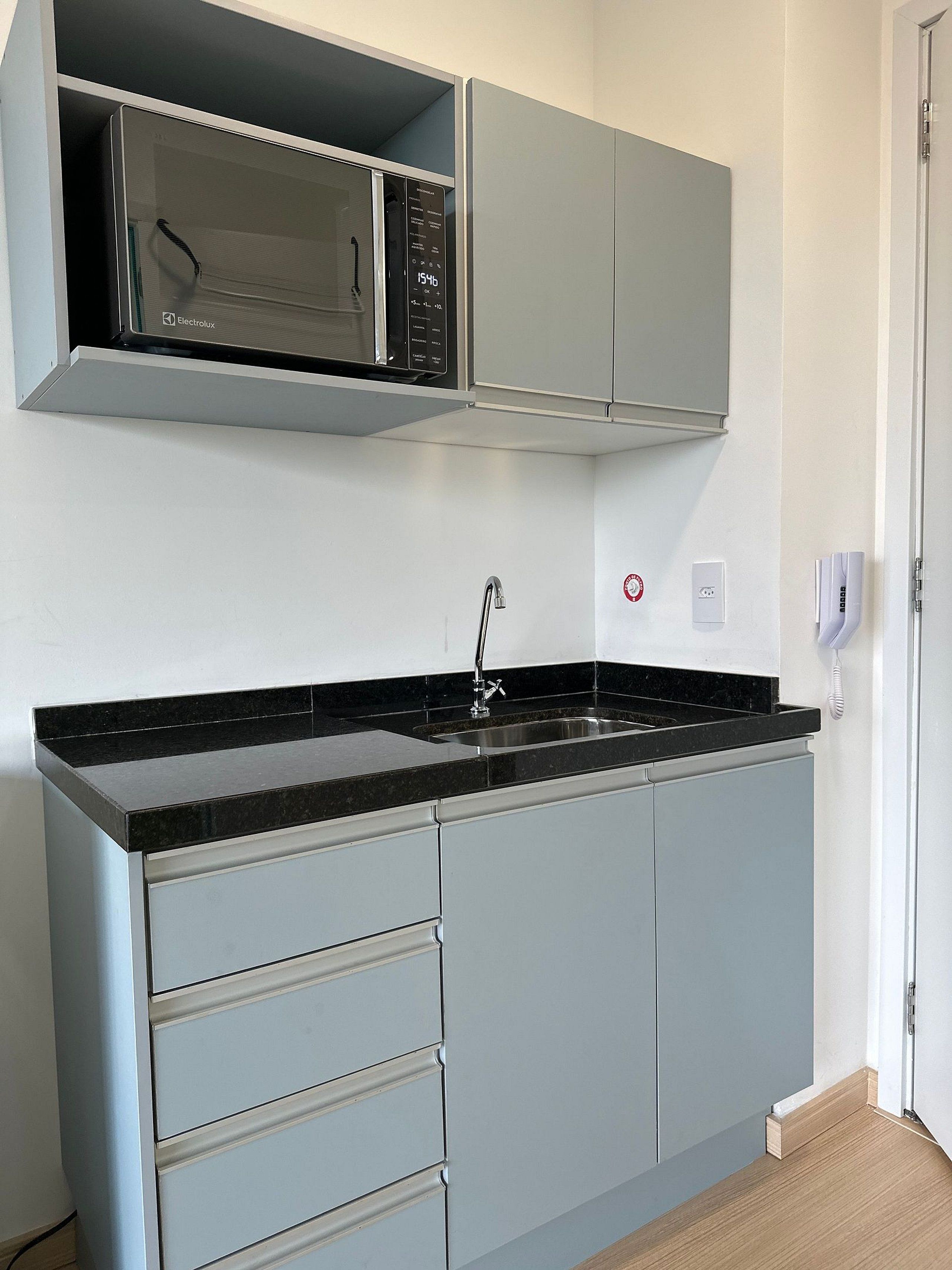 A kitchen with a black countertop, grey cabinets, a stainless steel sink, and a microwave.