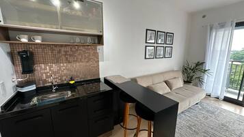 A modern kitchen with black cabinets, a black countertop, and a glass backsplash. There is a bar counter with stools, a beige sofa, and a potted plant.