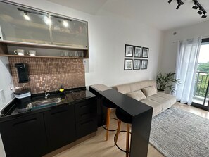 A modern kitchen with black cabinets, a black countertop, and a glass backsplash. There is a bar counter with stools, a beige sofa, and a potted plant. - TrevizZo Borba Gato (São Paulo)