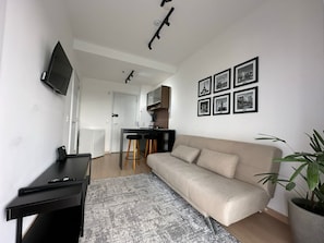 Una sala de estar moderna con un sofá, televisión, escritorio y una cocina al fondo.