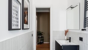 Un couloir étroit, avec un panneau encadré sur le mur de gauche, un pot de fleurs au centre, et une salle de bain moderne avec un lavabo et un miroir sur le côté droit.