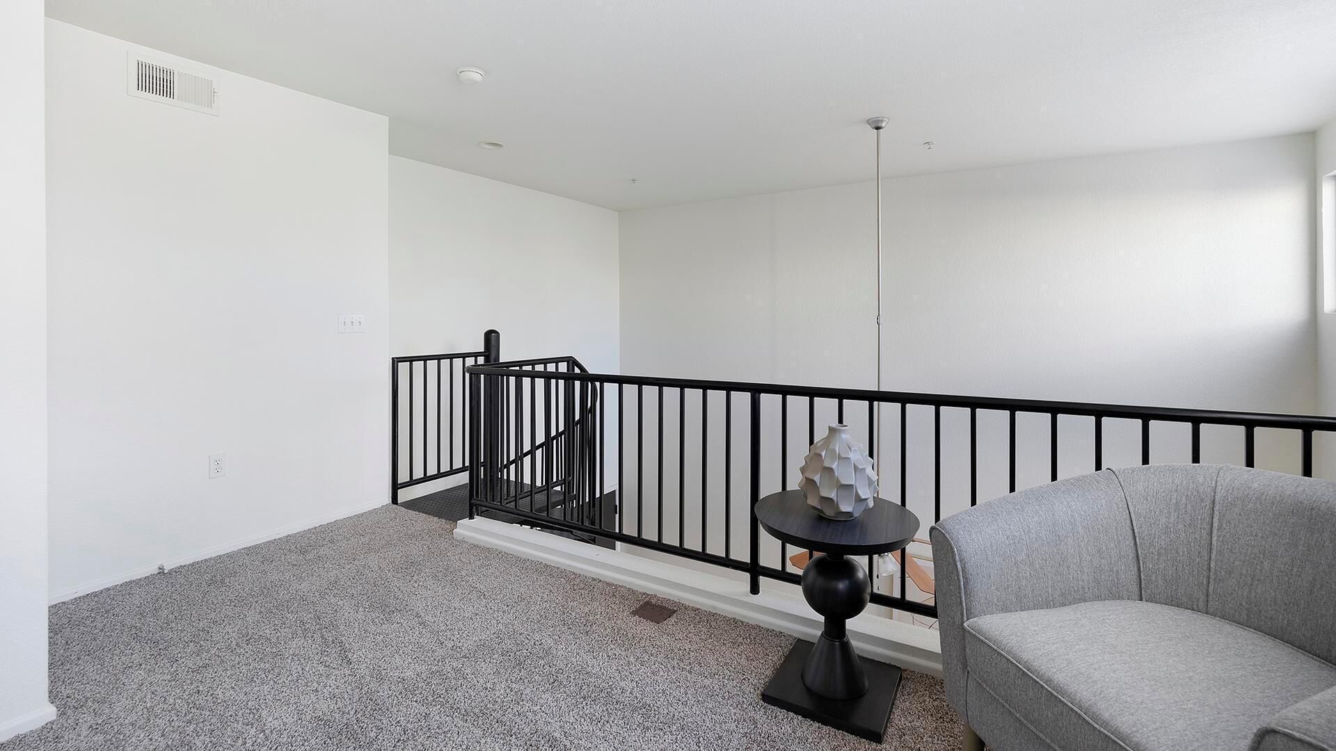 A modern apartment with a grey carpet, a grey sofa, a black railing, and a small table with a white vase.