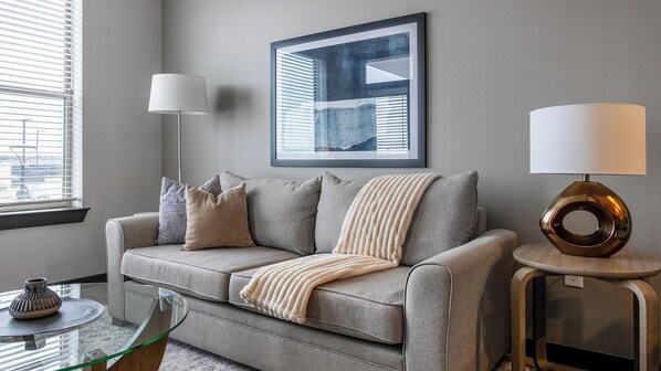 A modern living room with a grey sofa, a glass coffee table, a framed artwork on the wall, and two floor lamps. - Landing | Stunning 1BD, Gym, Clubhouse (Kyle)