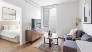 Ein modernes Schlafzimmer mit einem Bett, einem Fernseher auf einer Holzkommode, einem kleinen runden Tisch mit einer Pflanze und einem grauen Sofa.