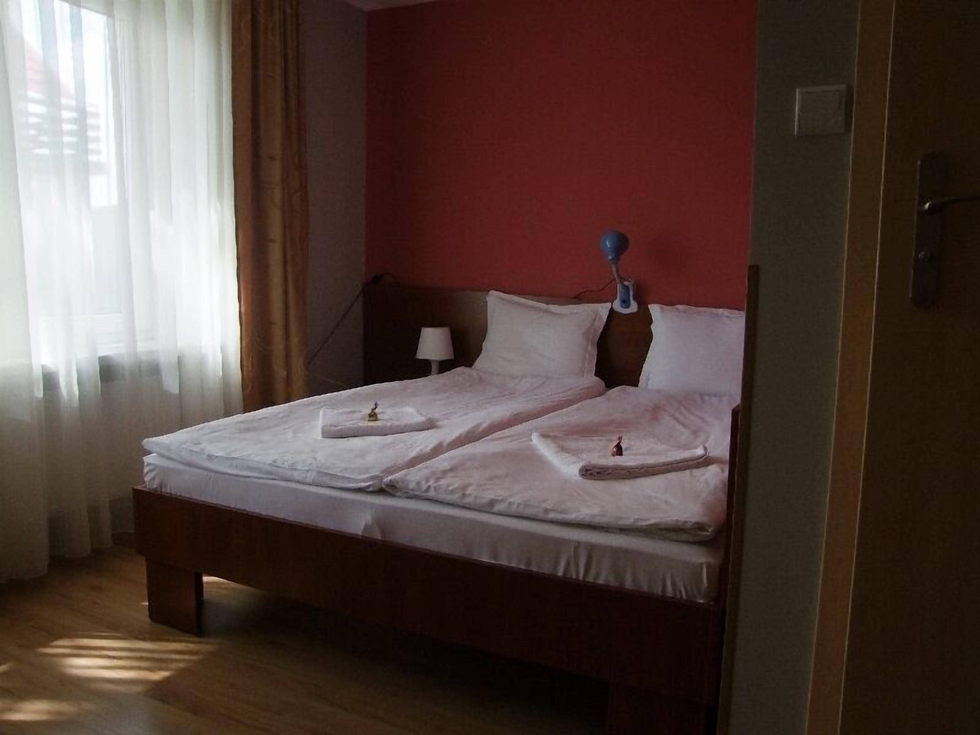 A neatly made double bed with pillows and a small bedside table, a lamp, and a window with sheer curtains.