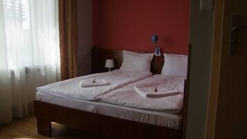 A neatly made double bed with pillows and a small bedside table, a lamp, and a window with sheer curtains.