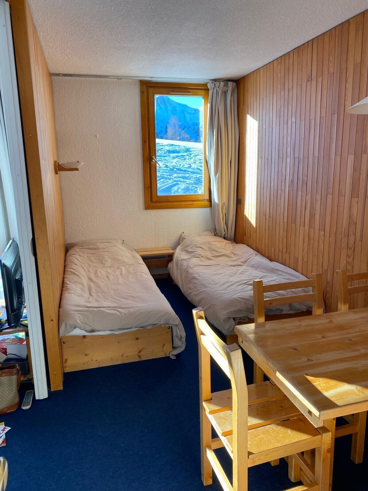 A small, compact room with two wooden beds, a wooden table, and a window with a view of a snowy landscape.