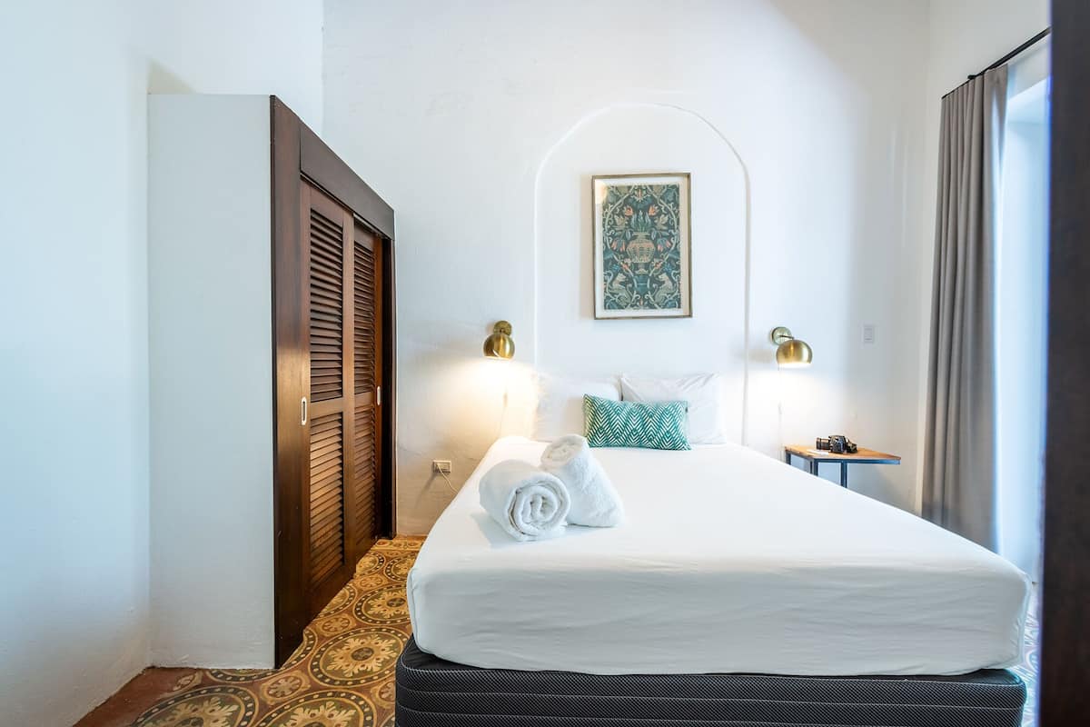 A hotel room with a bed, two bedside lamps, a wall-mounted picture, and a window with curtains.