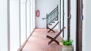 Un pasillo con piso de baldosas, paredes blancas y techo de madera. Se observan puertas a la derecha, una escalera con barandilla y una planta en el suelo.