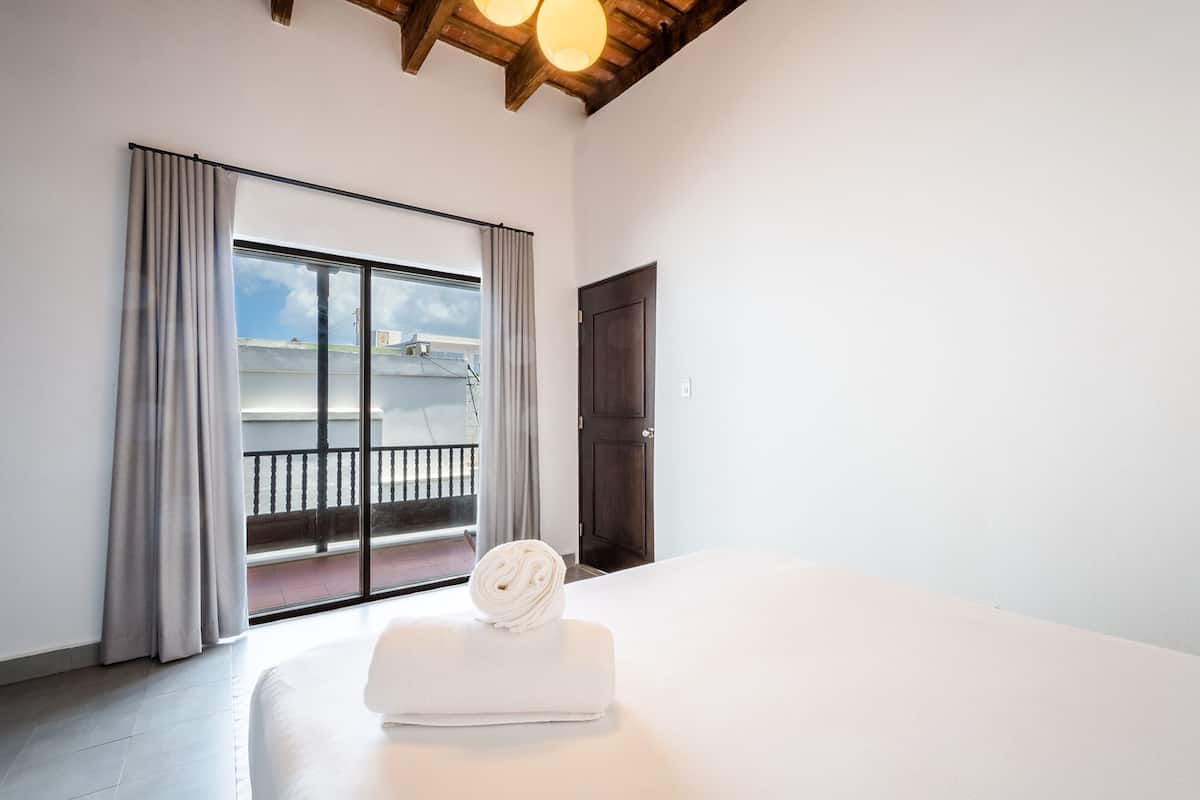 A hotel room with a large sliding glass door leading to a balcony, a white bed, and a dark wooden door.