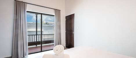 Habitación de hotel con un amplio ventanal corredera que da a un balcón, una cama blanca y una puerta de madera oscura.