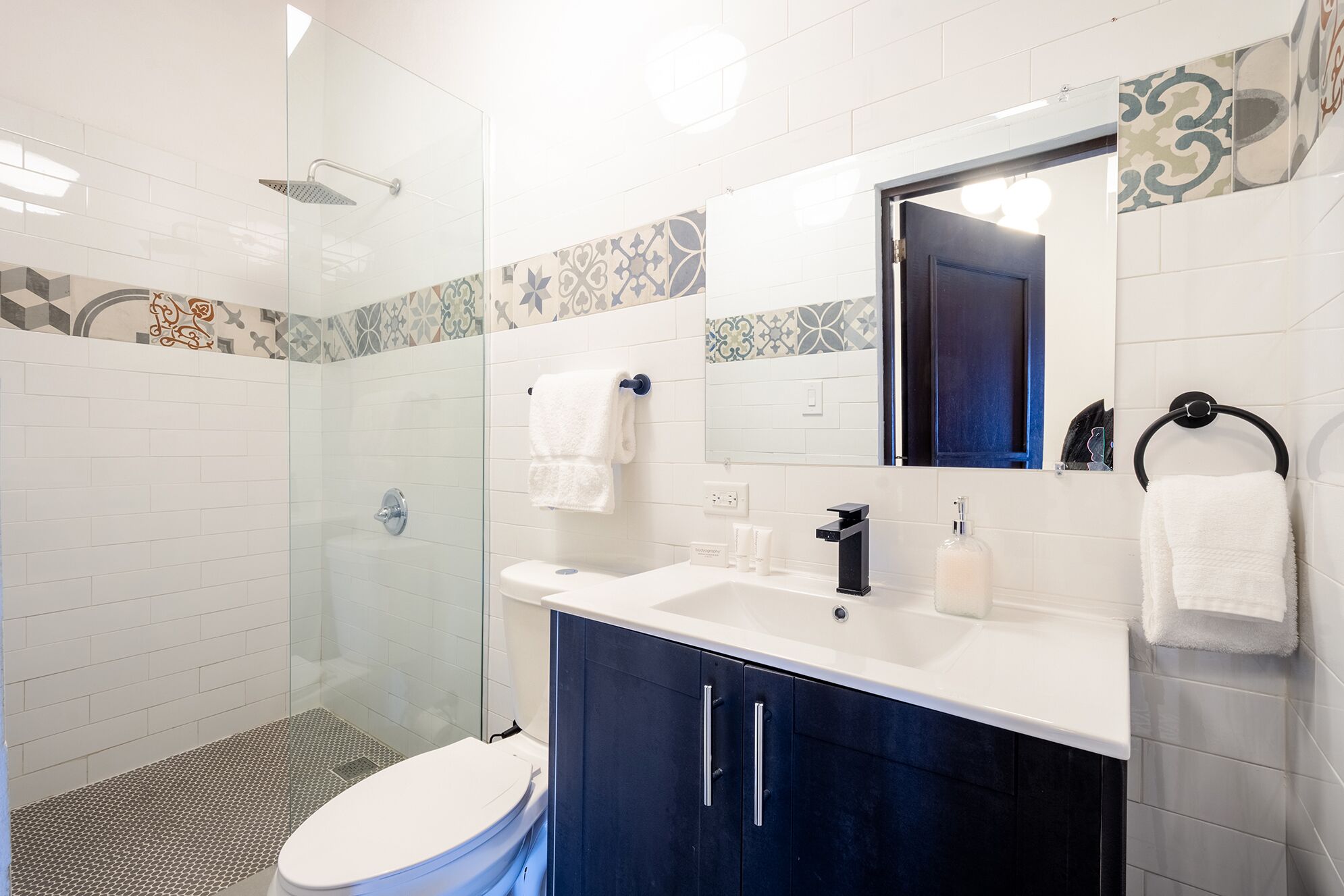 A modern bathroom with a glass shower enclosure, a white toilet, a vanity with a black faucet, and a mirror with a decorative tile backsplash.