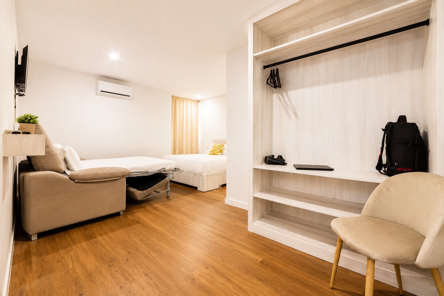 A compact room with a sofa, a bed, a chair, and a built-in wardrobe with shelves.