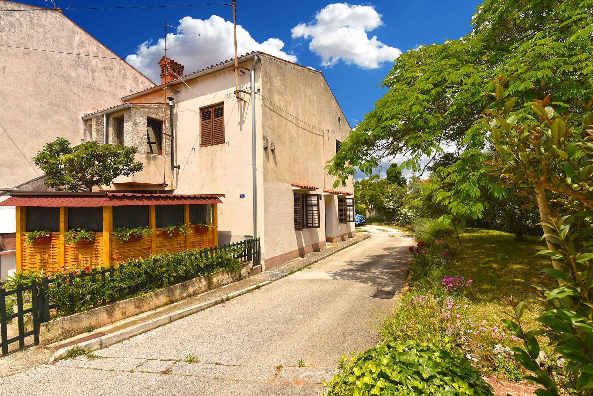 Una calle estrecha con un edificio que tiene un balcón con plantas, un pequeño jardín y un cielo azul despejado.