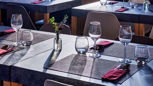 A dining area with tables set for two, each with a vase of flowers, wine glasses, and cutlery. A large window offers a view of the sky and distant landscape.