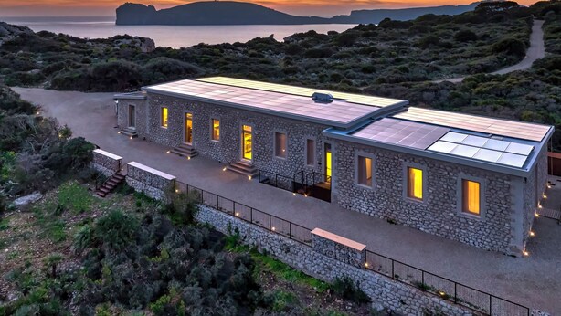 A stone building with multiple windows and a flat roof, surrounded by natural landscape and a body of water.