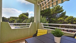 A balcony with a table and chairs, a potted plant, and a view of trees and parked cars. - Holiday apartment with air conditioning (Pula)