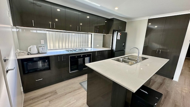 A modern kitchen with dark cabinetry, a central island with a sink, built-in ovens, and a window with blinds.