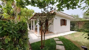 A small white building with a tiled roof, a covered patio area, and a well-maintained garden with a lawn and a hedge. - Holiday home mit Terrasse und Klimaanlage (Santa Maria di Castellabate)
