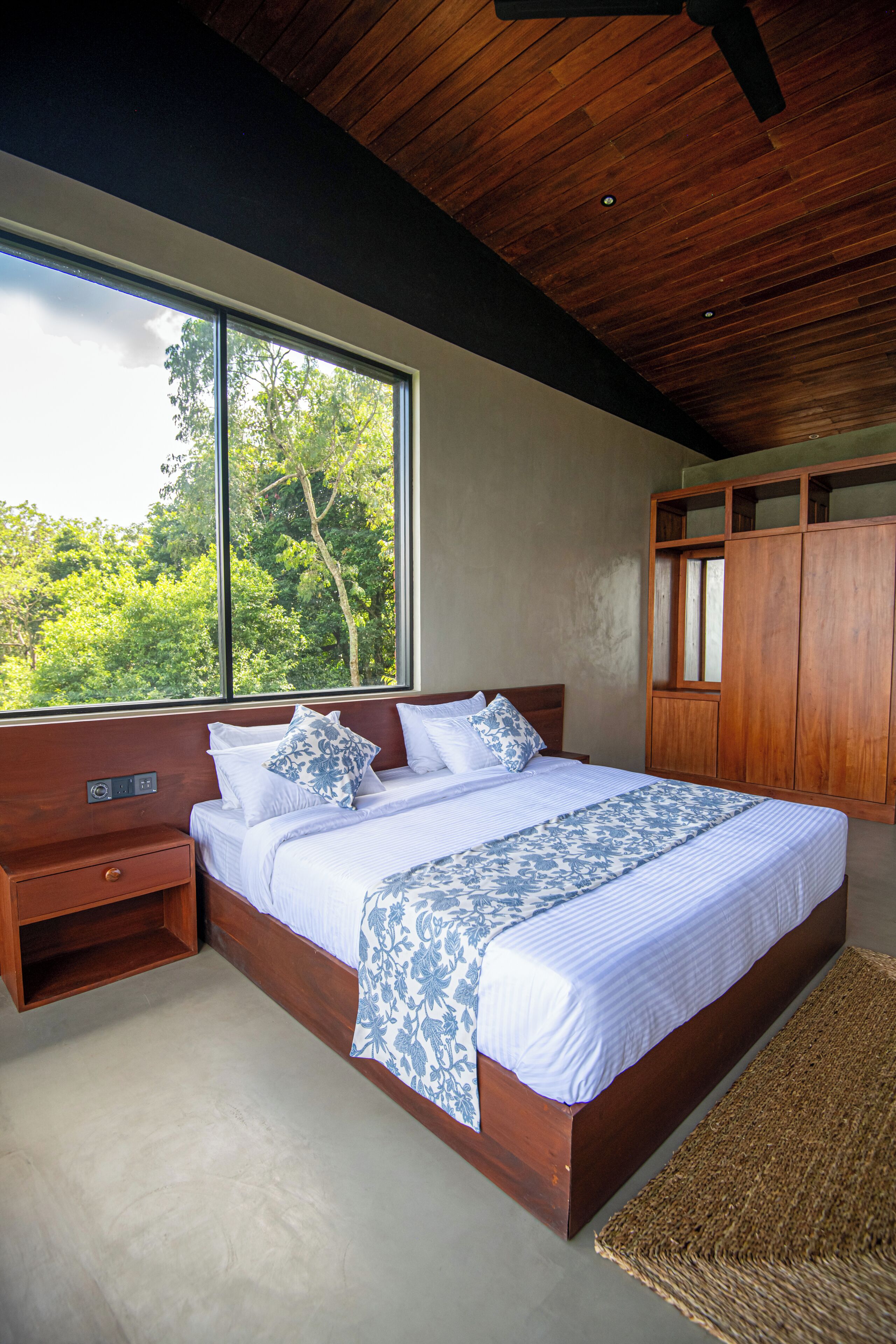 A bedroom with a large bed, wooden headboard, a nightstand, a wardrobe, and a view of trees through a window.