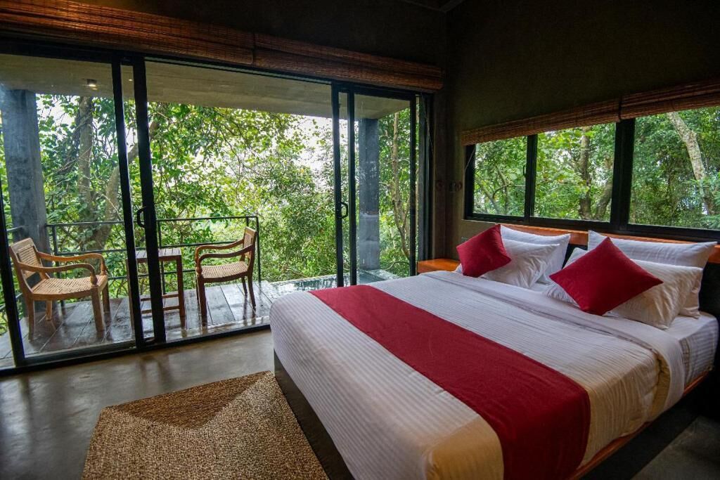 A large bed with red and white bedding, a wooden headboard, and a view of lush greenery through large windows.