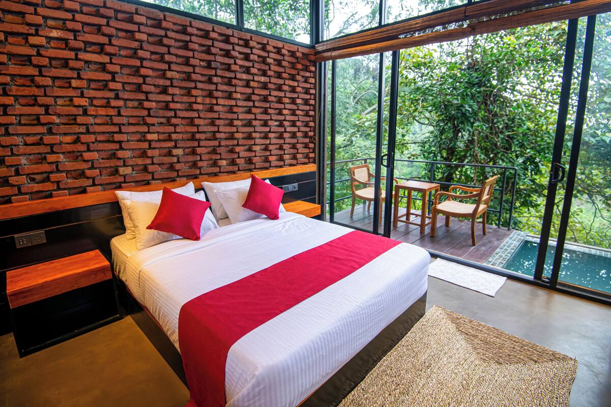 Ein modernes Hotelzimmer mit einer Ziegelwand, einem großen Bett mit rot-weißer Bettwäsche, einem Balkon mit Tisch und Stühlen sowie Blick auf üppiges Grün.