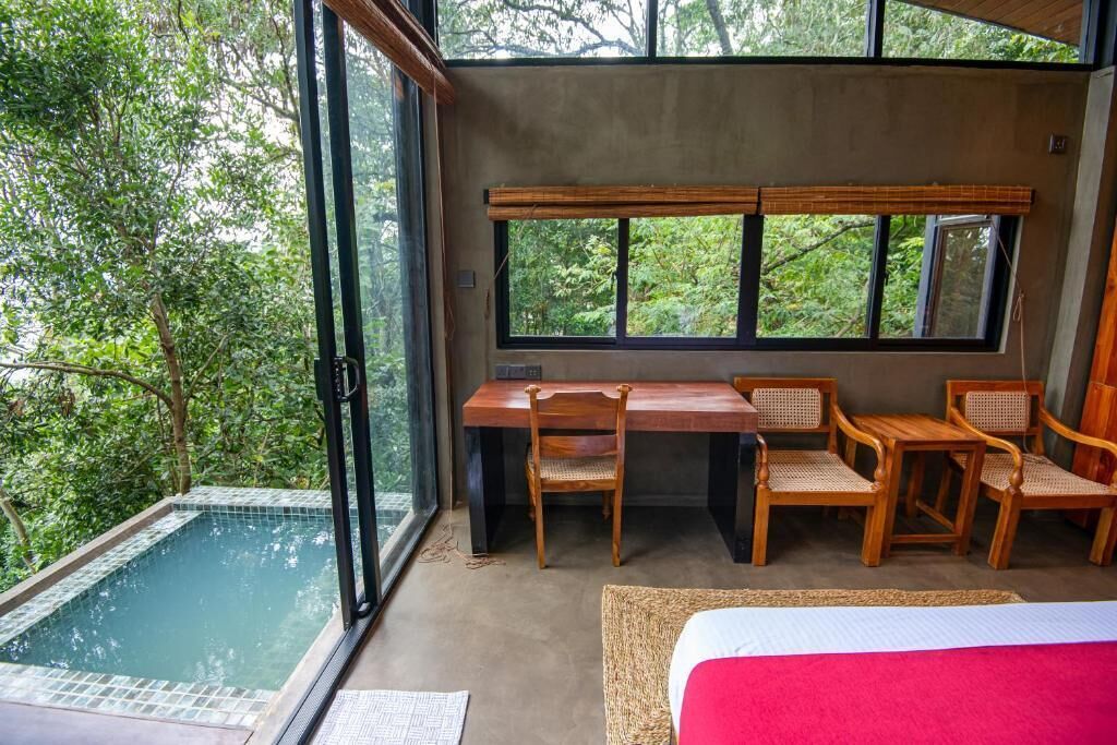 A room with a glass wall, a pool, a wooden desk with chairs, and a bed.
