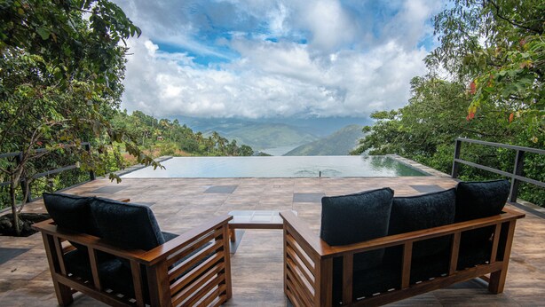 A wooden bench with cushions facing a pool, surrounded by lush greenery and a mountainous backdrop. - Nuga Retreat (Illagolla)