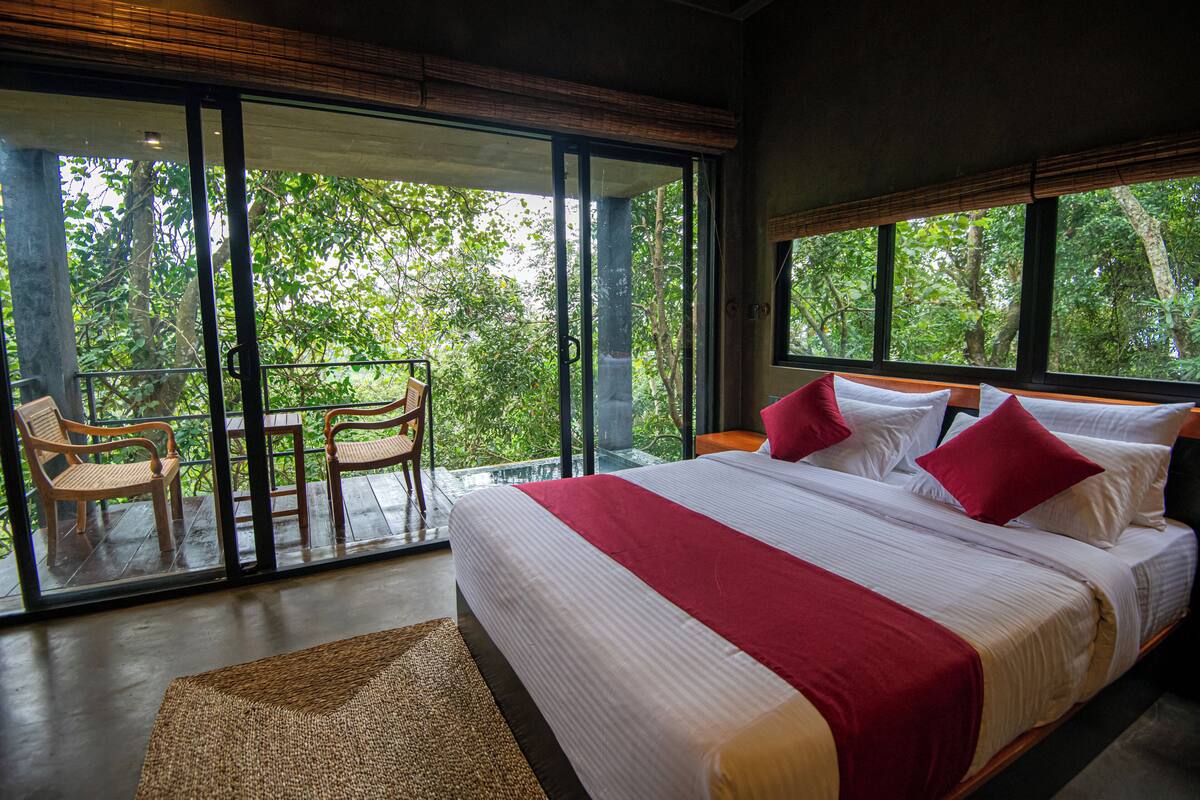 Ein großes Bett mit weißer und roter Bettwäsche, ein hölzerner Kopfteil und Blick auf üppiges Grün durch große Fenster.
