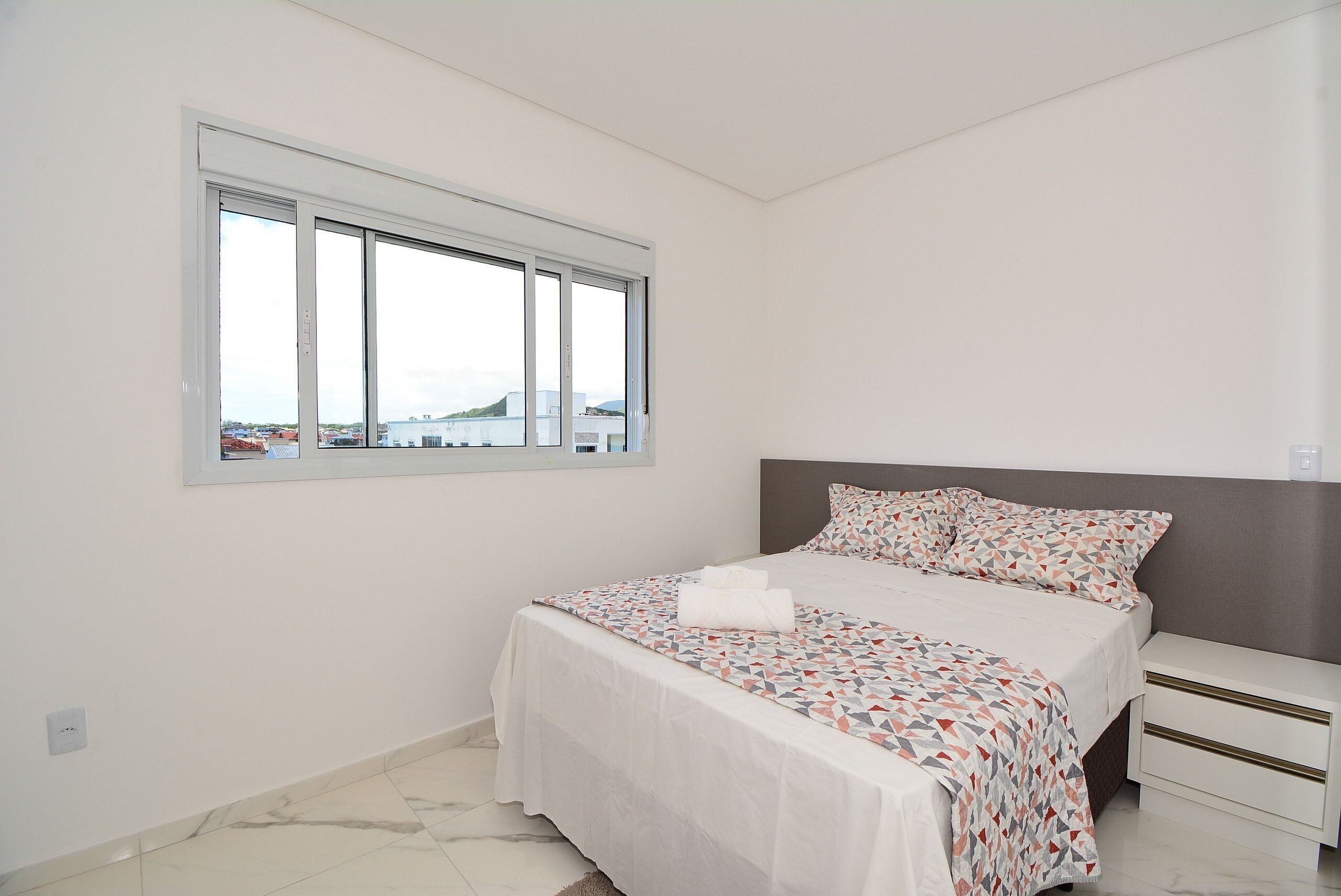 Un dormitorio con un ventanal grande, una cama con ropa de cama estampada y una mesita de noche.