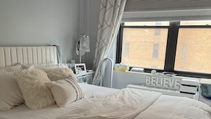 Una cama bien tendida con cabecera blanca, una mesita de noche con lámpara y una ventana que da a un edificio de ladrillo.