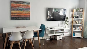 Una sala de estar moderna con un televisor de pantalla plana, un cuadro abstracto colorido, un escritorio blanco con sillas, una estantería de madera y una pequeña planta en maceta.