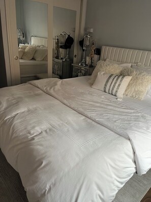 A neatly made bed with a white comforter and pillows, a bedside table with a lamp, and a mirror reflecting the room.