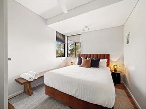 Une chambre moderne avec un grand lit, un banc avec des serviettes, une table de chevet avec une lampe et une fenêtre avec des stores.