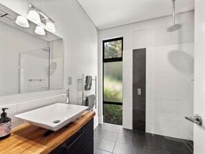 Une salle de bain moderne avec un grand miroir, un meuble-lavabo en bois, une douche avec une porte en verre et une vue sur la verdure à l’extérieur.