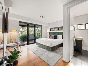 Une chambre moderne avec un grand lit, un coin salon avec des fauteuils, une vue à travers une porte vitrée et une salle de bain avec un lavabo et un miroir.