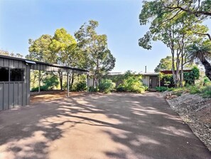 A paved driveway leading to a building with a covered structure on the left and a small house on the right, surrounded by trees and vegetation.