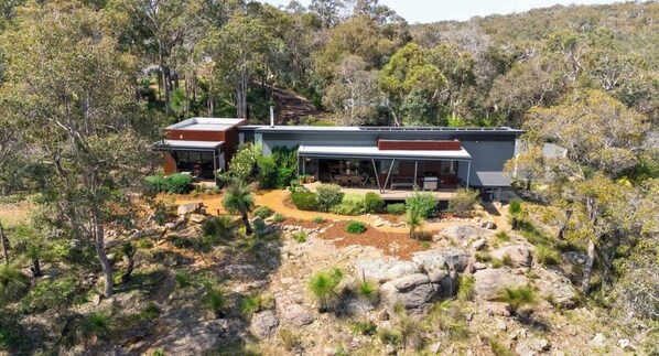 Une maison moderne dotée d’une grande terrasse, entourée d’une végétation luxuriante et d’un paysage rocheux.