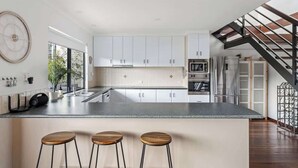 A modern kitchen with white cabinets, a central island with stools, a large clock on the wall, and a staircase leading to another level. - Arbre Retreat—Serene Bushland Escape Near Beach (Dunsborough)