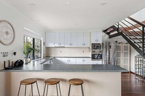 Una cocina moderna con armarios blancos, una isla central con taburetes, un reloj grande en la pared y una escalera que conduce a otro nivel.