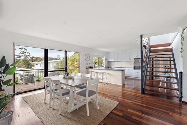 Una cocina moderna con una isla central, una zona de comedor con mesa y sillas, y una escalera que sube a un nivel superior.